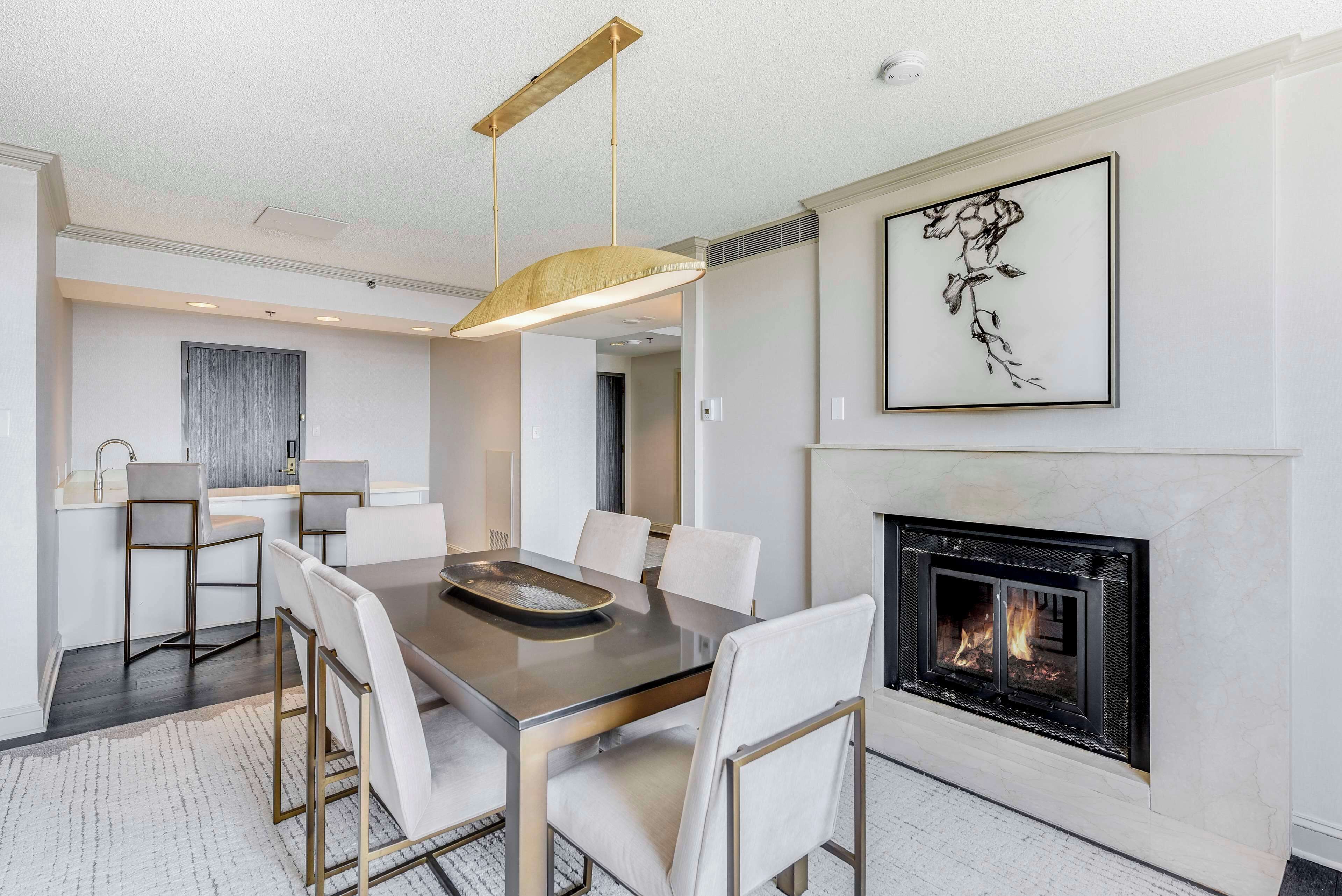 A modern dining area with a glass table, white chairs, a pendant light, and a fireplace.