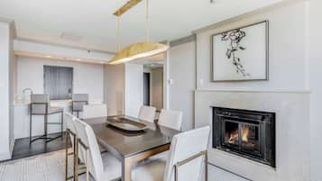 A modern dining area with a glass table, white chairs, a pendant light, and a fireplace.