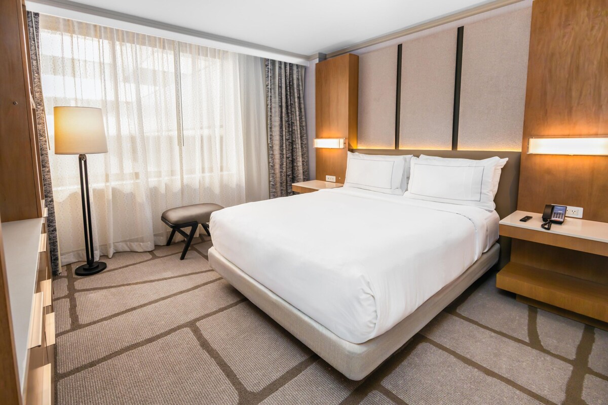 A hotel room with a large bed, a bedside table with a lamp, a chair, and a desk with a phone.