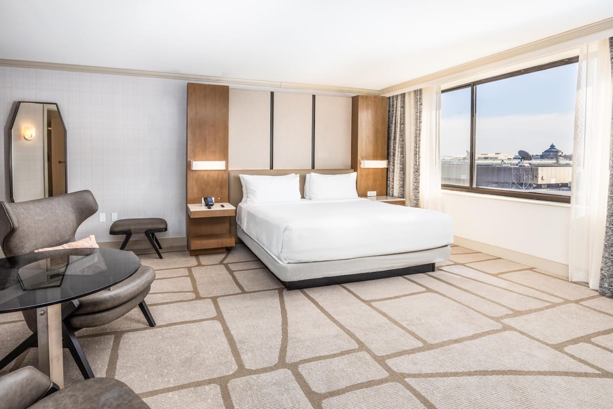 A hotel room with a bed, a desk, a chair, a mirror, and a large window with a city view.
