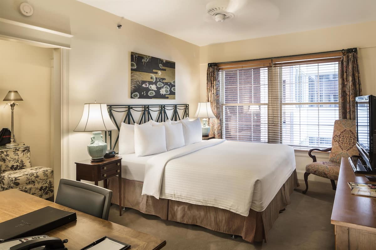 A hotel room with a large bed, two bedside tables with lamps, a chair, a desk, and a window with blinds.