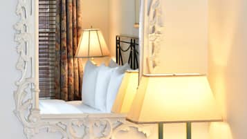 A hotel room with a large ornate mirror, a wooden desk, a black chair, and a bed with white pillows and a floral curtain.