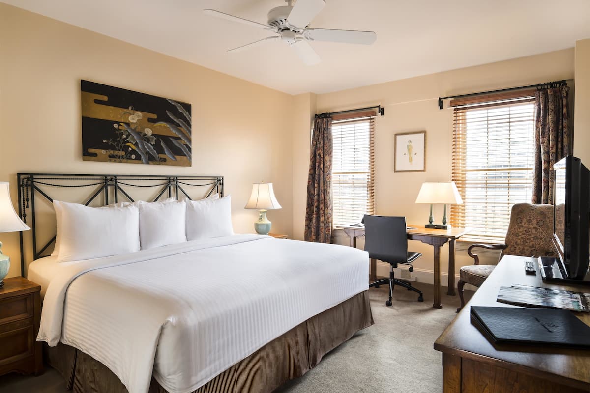 A hotel room with a large bed, two bedside tables, a desk with a chair, a television, and a ceiling fan.