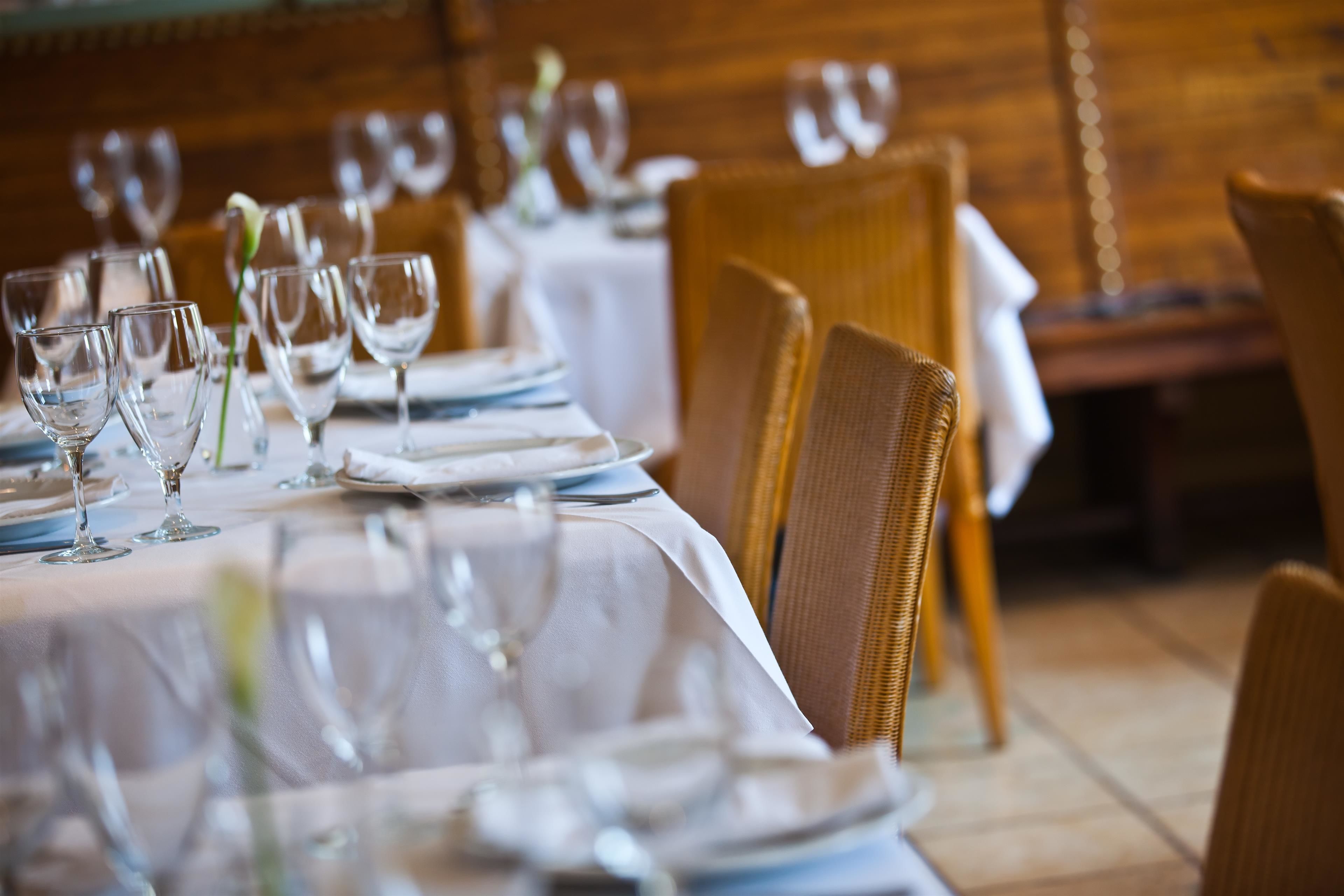 A dining table set with wine glasses, plates, and cutlery in a restaurant with wooden chairs and a white tablecloth.
