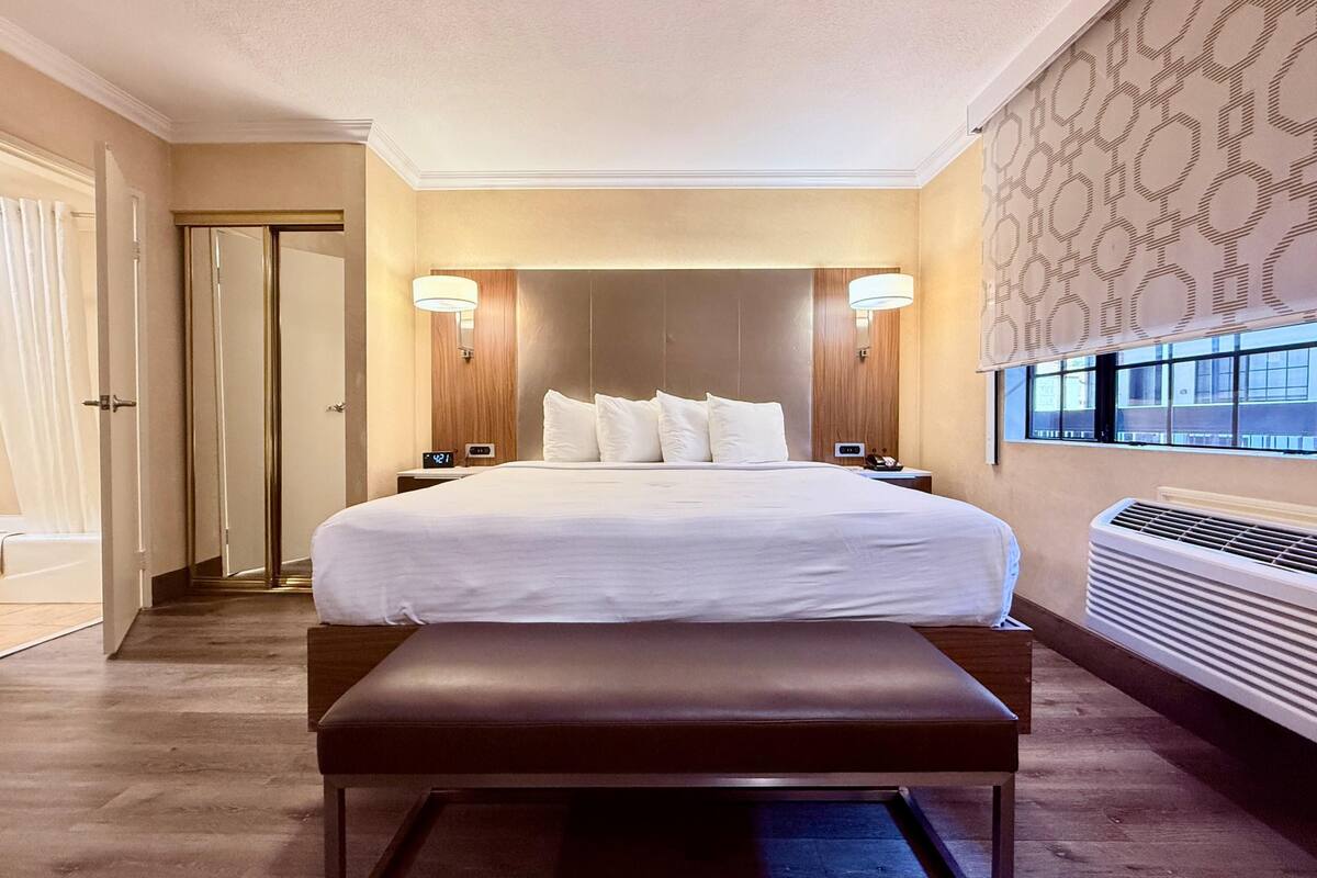 Ein Hotelzimmer mit einem großen Bett, Nachttischlampen, einer Bank, einer Tür und einem Fenster mit Blick.