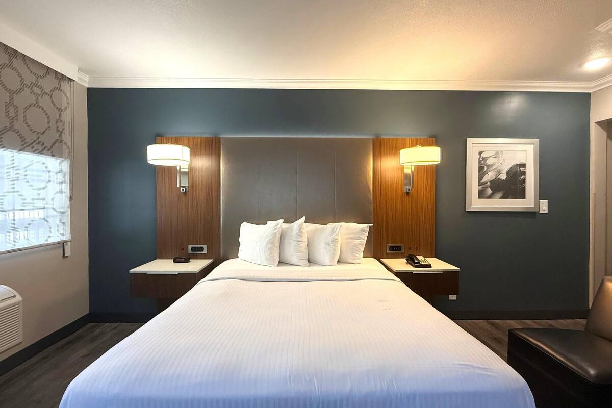 Ein Hotelzimmer mit einem großen Bett, zwei Nachttischen, einer Wandlampe, einem gerahmten Bild und einem Fenster mit Jalousien.
