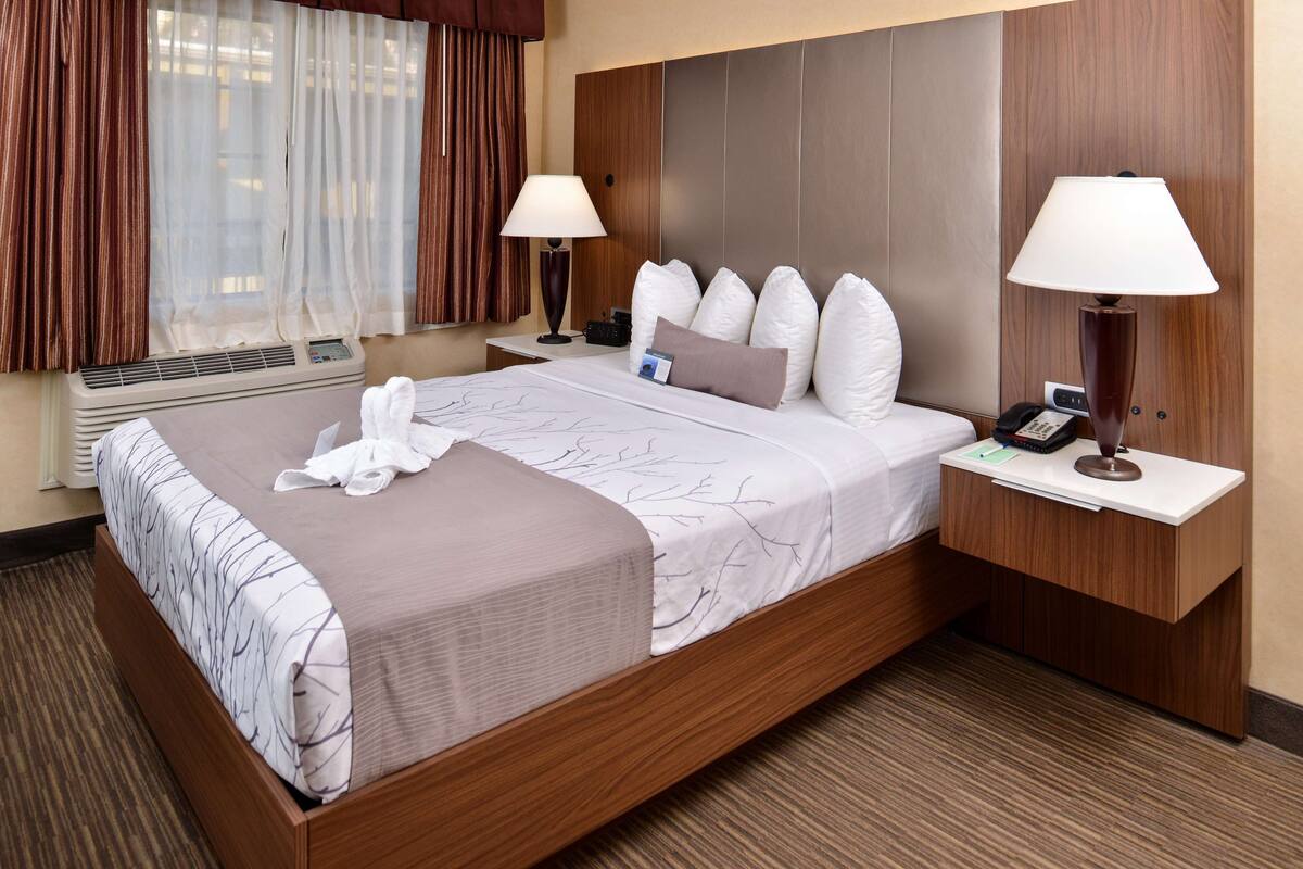 Ein Hotelzimmer mit einem großen Bett, zwei Nachttischen, einer Wand-Klimaanlage und einem fensterbepflanzten Fenster mit Vorhängen.