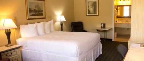 Une chambre d’hôtel avec un grand lit, deux tables de chevet avec des lampes, une chaise, un bureau avec un miroir et une salle de bain avec un lavabo et un miroir.