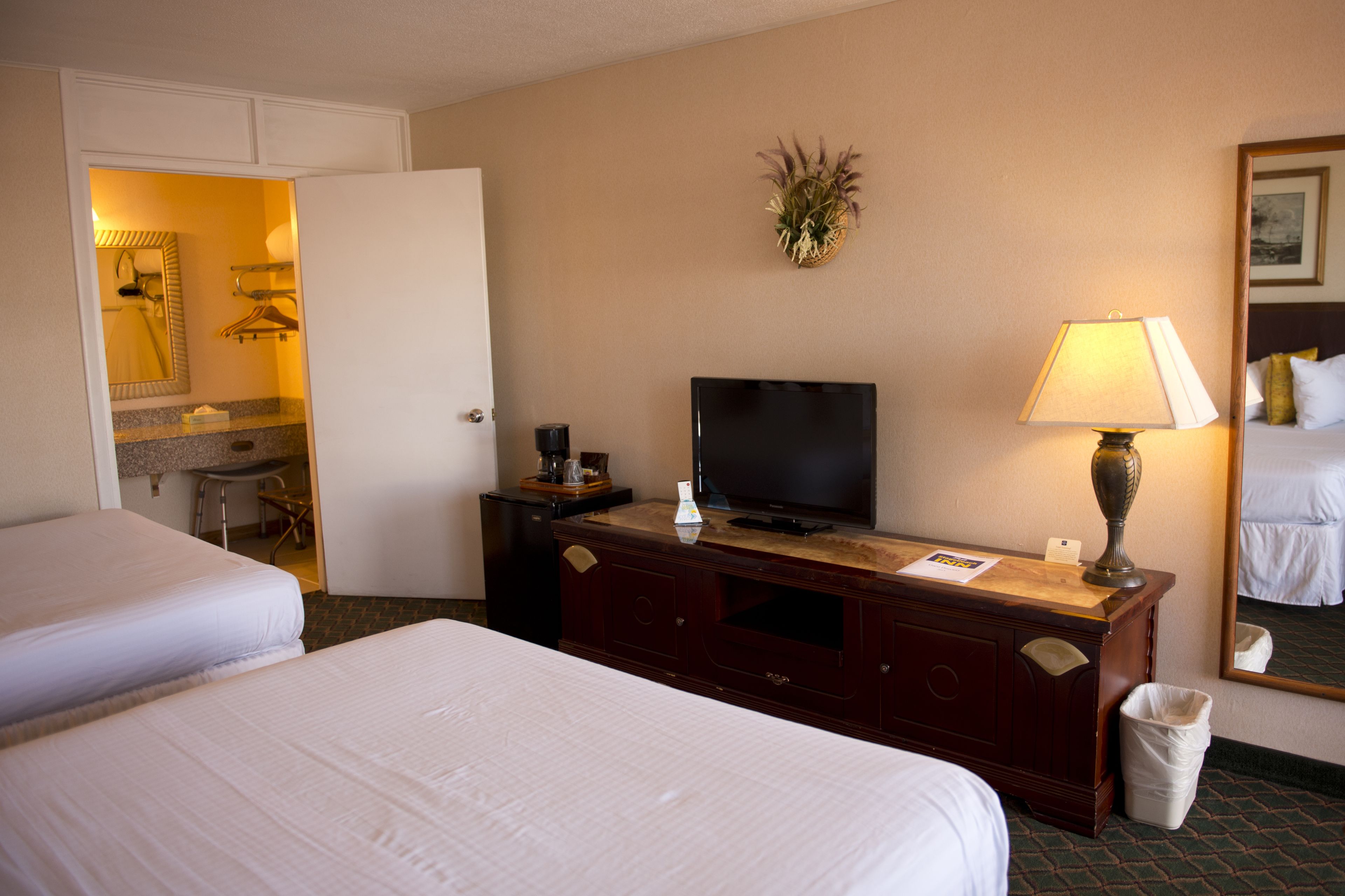 A hotel room with two beds, a TV, a lamp, a mirror, and a small desk with a coffee maker.