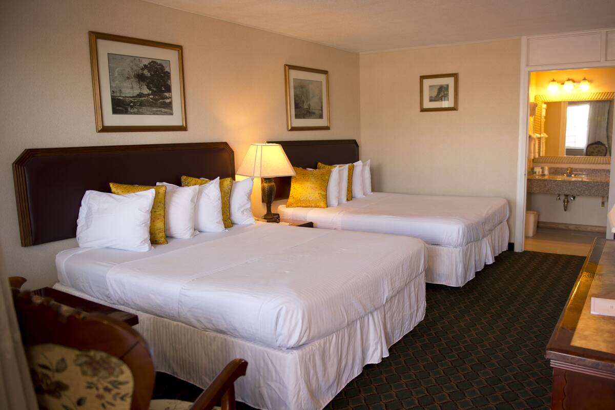 Une chambre d’hôtel avec deux lits, un bureau, une chaise, une lampe et une salle de bain équipée d’un lavabo et d’un miroir.