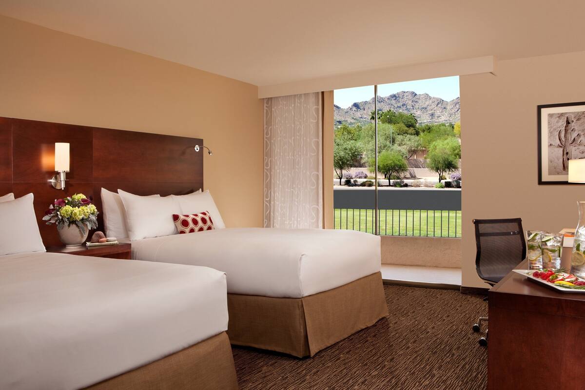 A hotel room with two beds, a desk, a chair, a large window with a view of trees and mountains, and a framed picture on the wall.