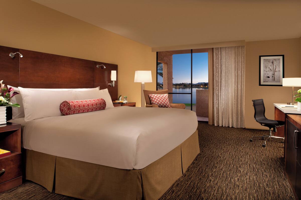 A large bed with a wooden headboard, a desk with a chair, and a view of the outdoors through a window.