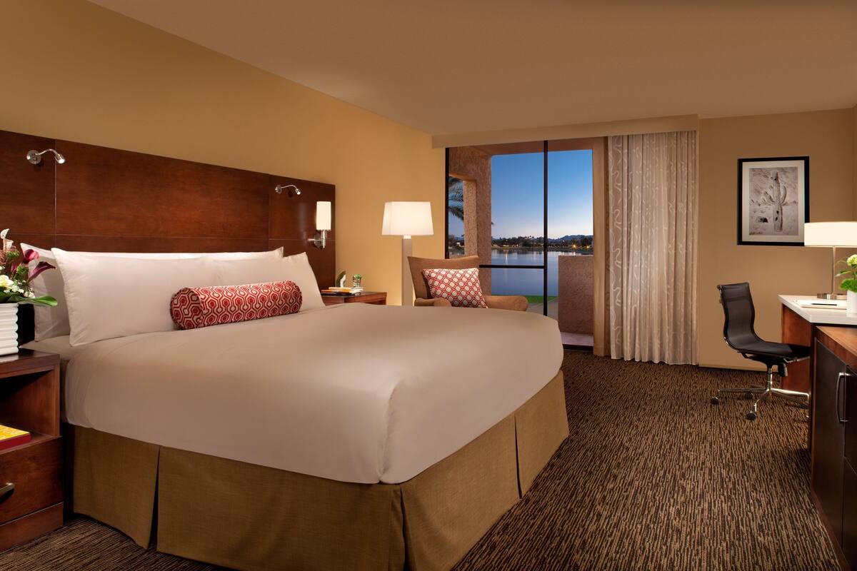 A hotel room with a large bed, a desk, a chair, a view of the outdoors, and a window with curtains.