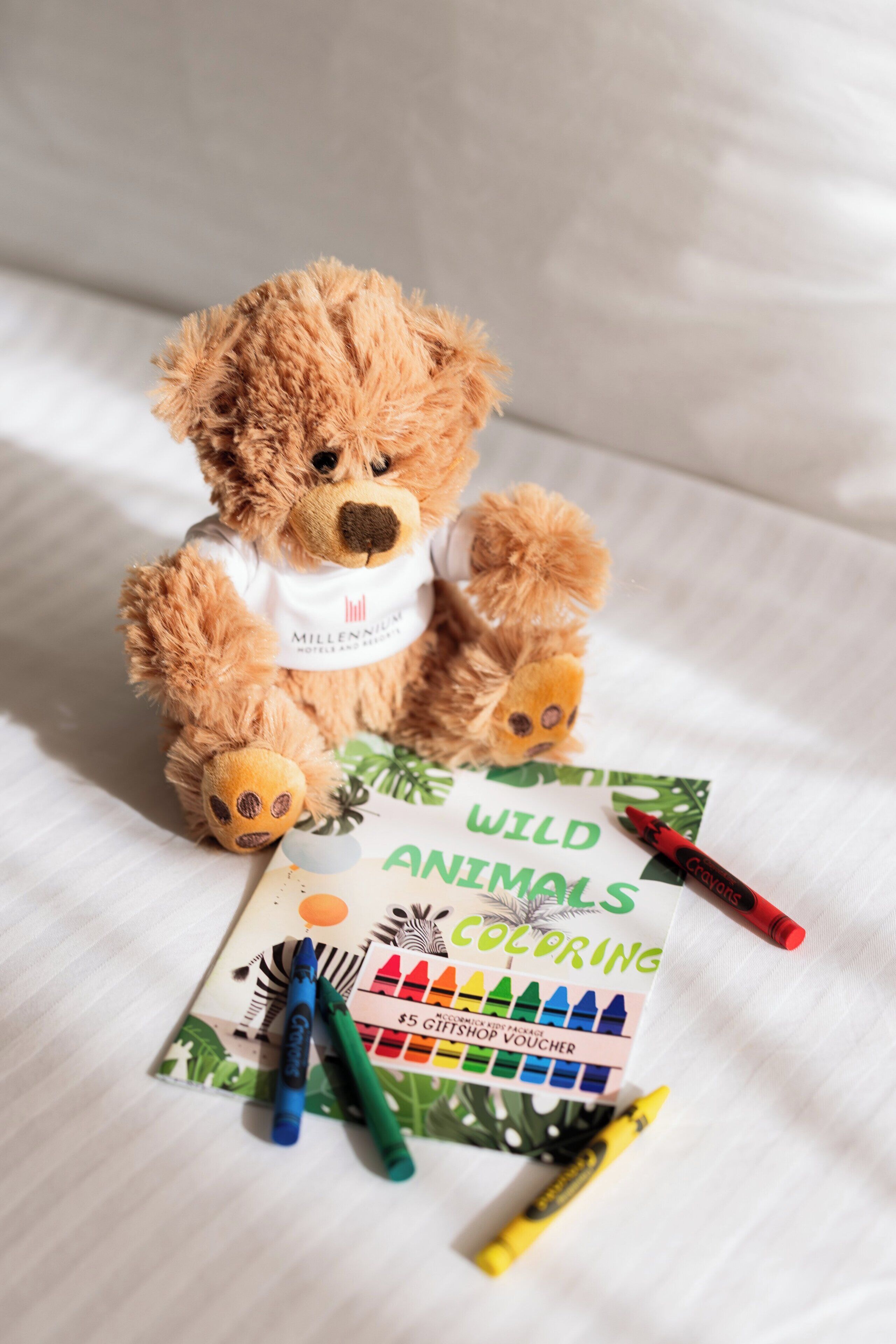 A teddy bear on a bed with a coloring book and crayons.