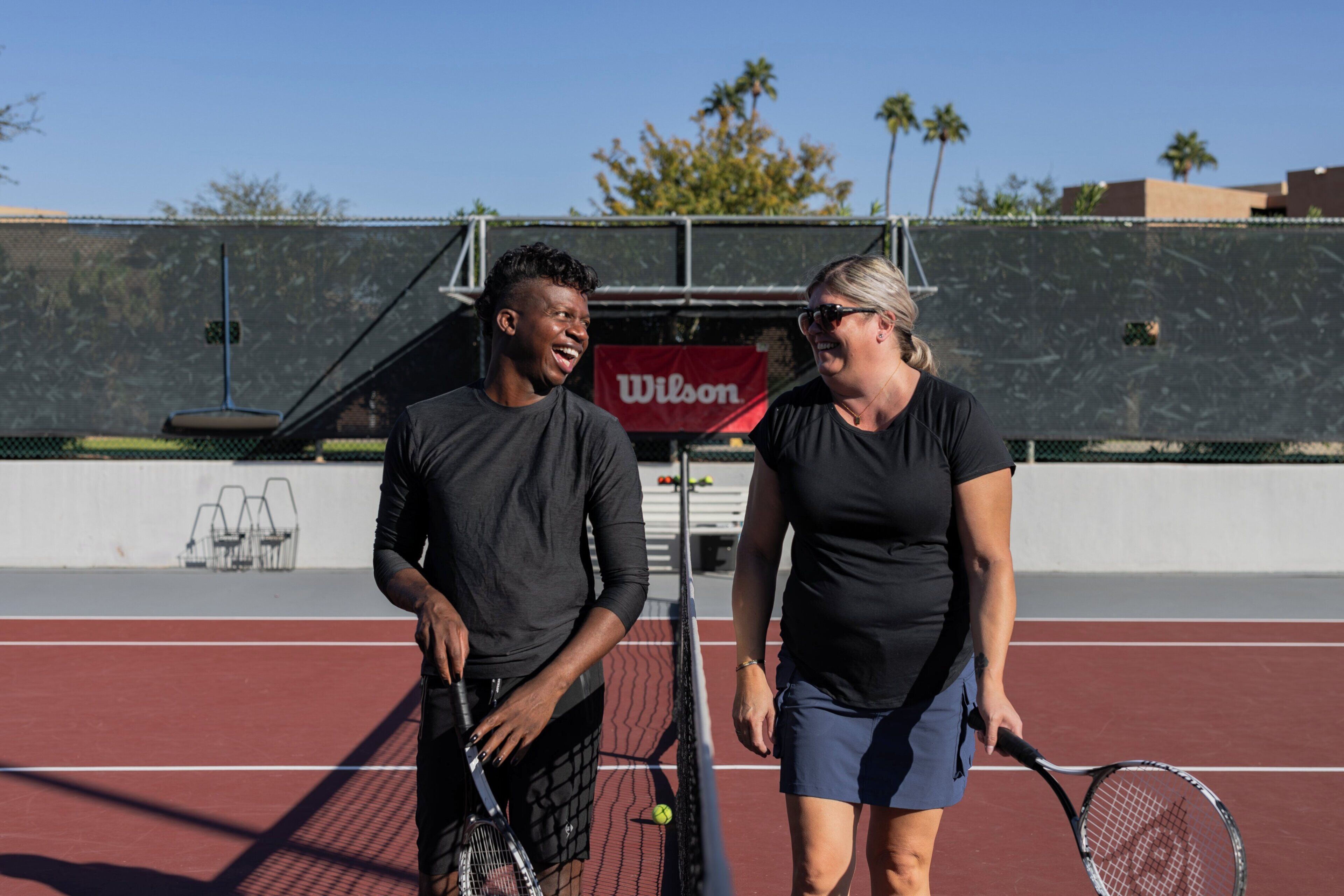 Two people on a tennis court with a fence and a 'Wilson' banner in the background.