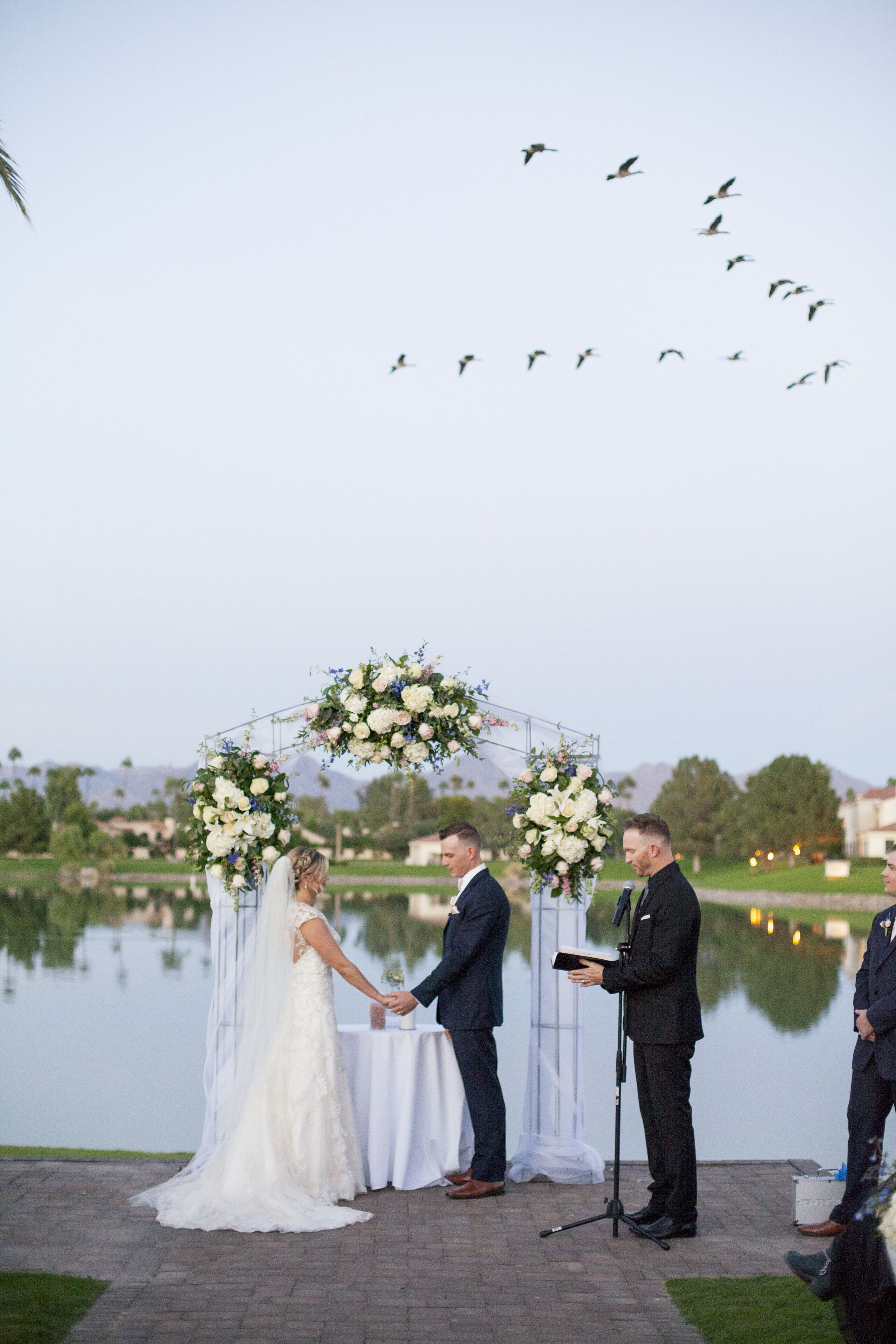 Eine Hochzeitszeremonie am See mit einem Blumenschmuck und Gästen im Hintergrund.