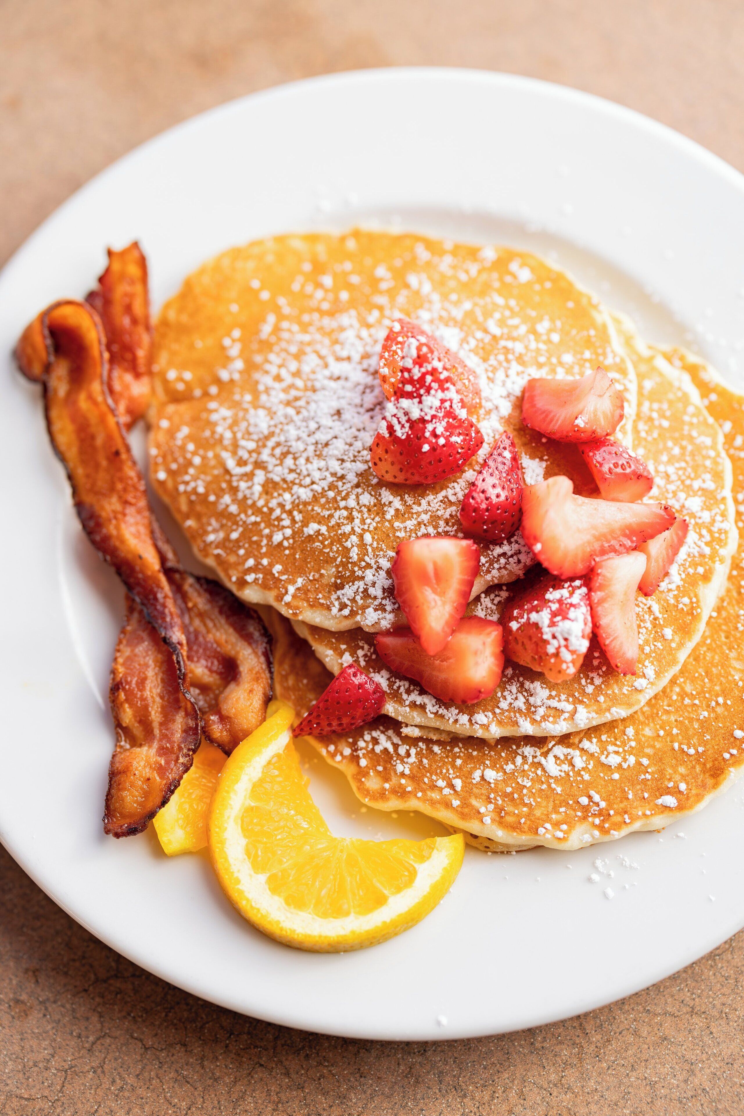 Un plato con tortitas, fresas, tocino y una rodaja de limón.