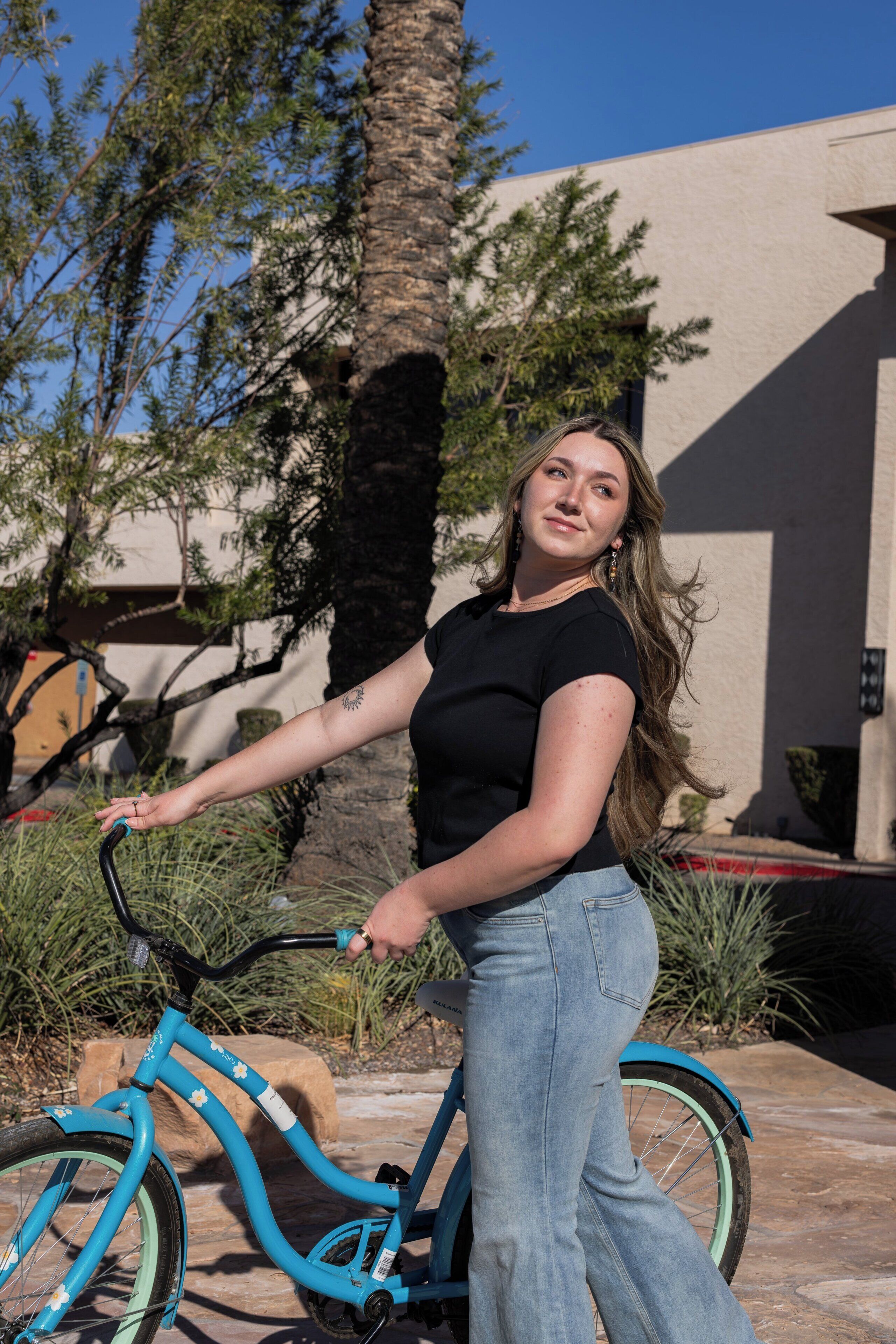 Une personne se tient à côté d’un vélo turquoise orné de motifs floraux, vêtue d’un haut noir et d’un jean bleu, dans un décor extérieur avec un bâtiment et des arbres en arrière-plan.