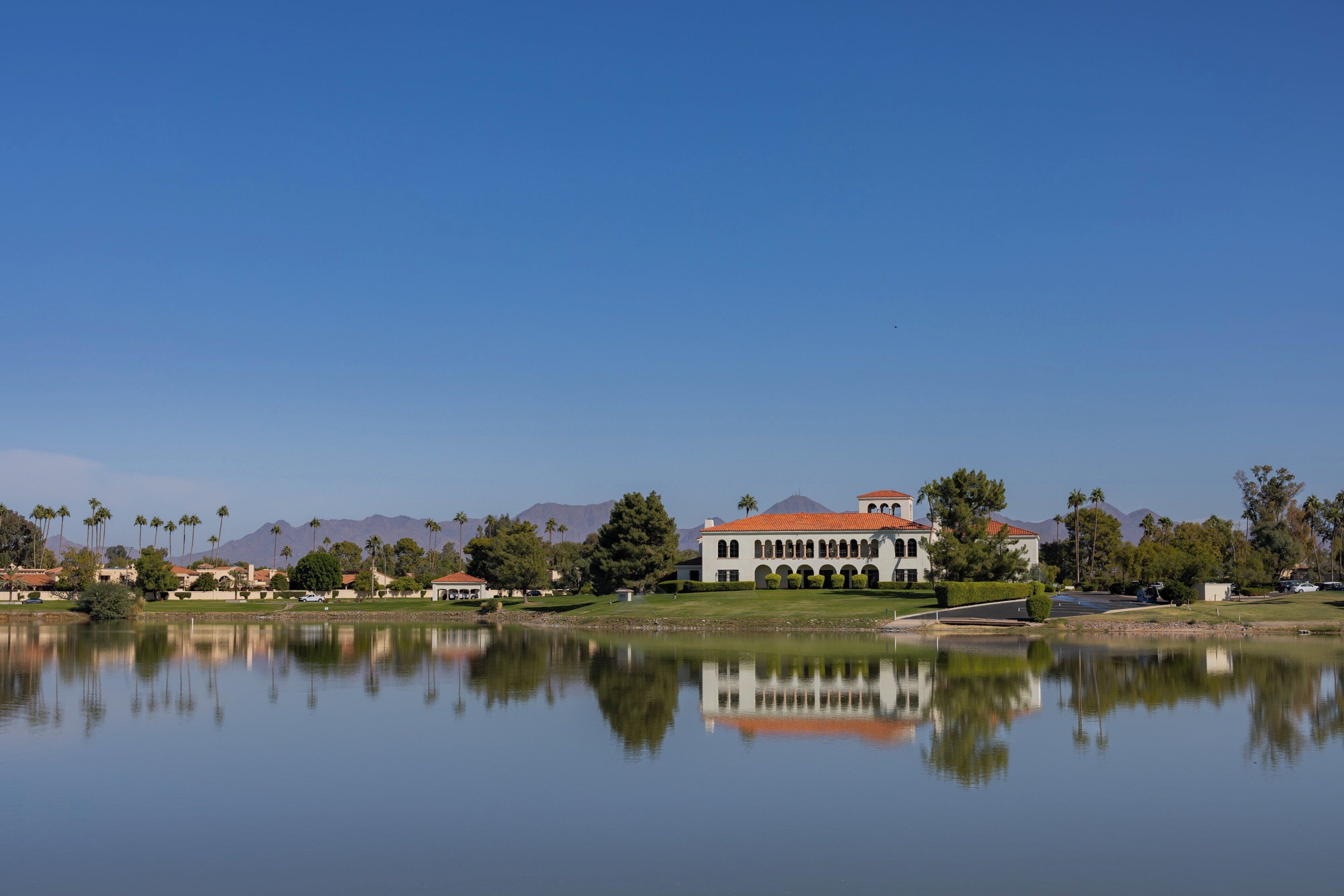 A large building with a red roof, a reflection in the water, and palm trees in the background.