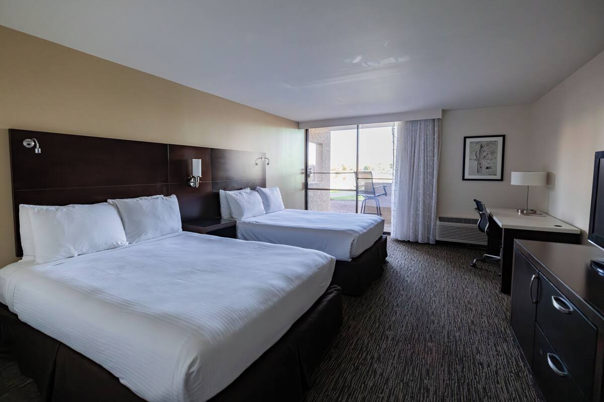 A hotel room with two beds, a desk, a chair, a lamp, and a window with a view.