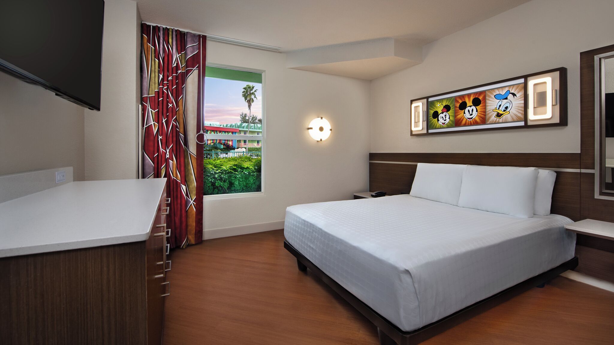 A hotel room with a bed, a desk, a TV, a window with curtains, and a wall with Mickey Mouse artwork.