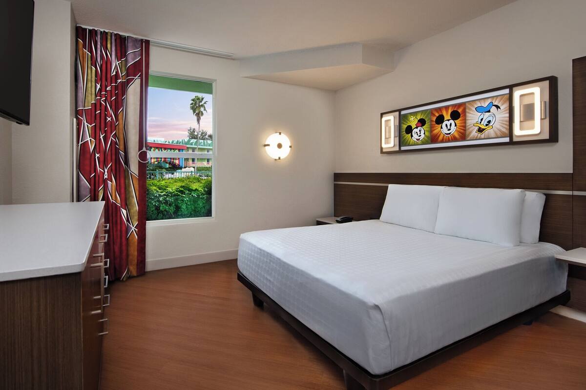 A hotel room with a bed, a desk, a TV, a window with curtains, and a wall with Mickey Mouse artwork.