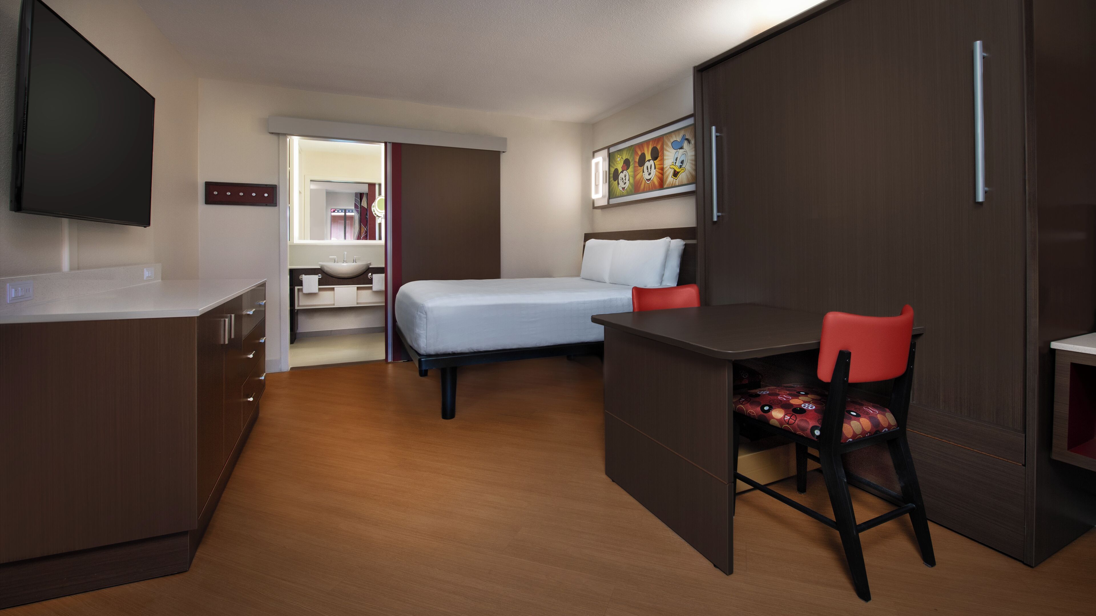 A compact hotel room with a bed, a desk, a chair, a TV, and a bathroom visible through an open door.