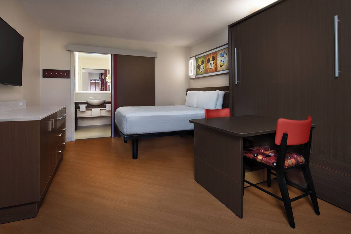 A compact hotel room with a bed, a desk, a chair, a TV, and a bathroom visible through an open door.