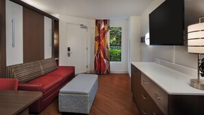 A modern hotel room with a red sofa, a small table, a bench, a TV, and a kitchen area with a white countertop and cabinets. - Disney's All-Star Music Resort (Lake Buena Vista)