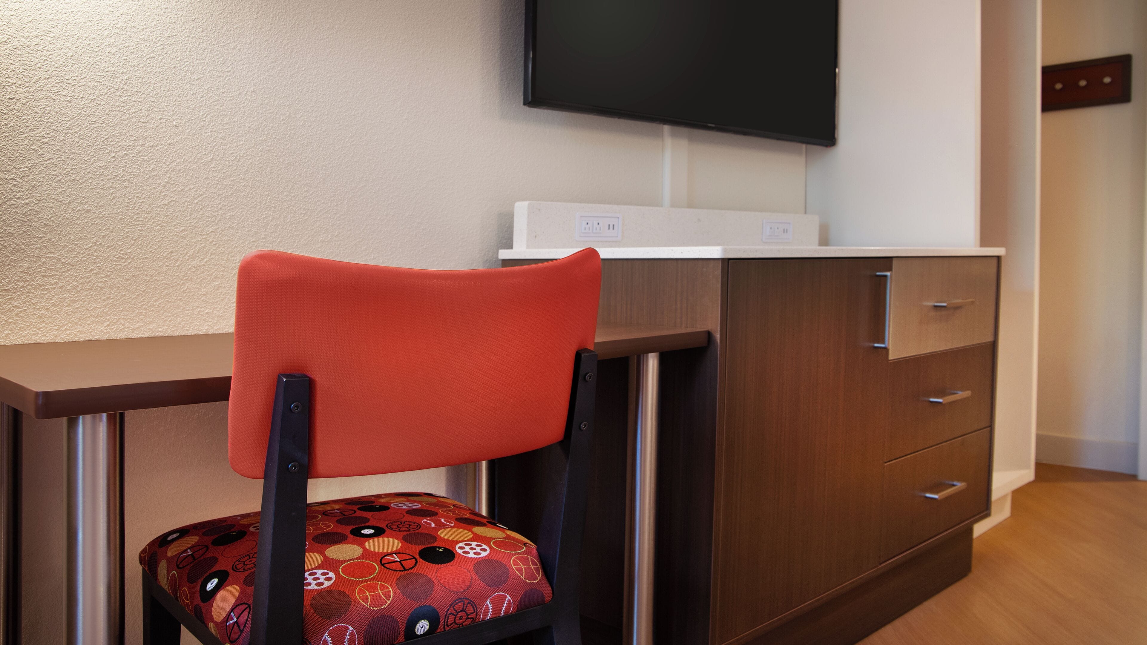 Une chambre moderne avec un bureau en bois, une chaise rouge agrémentée d’un coussin à motifs, un téléviseur à écran plat fixé au mur et une commode en bois avec des tiroirs.
