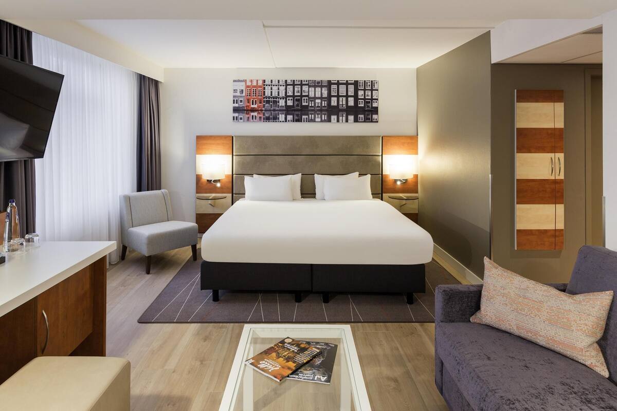Quarto de hotel com cama grande, escrivaninha, cadeira, sofá, televisão e uma obra de arte na parede.