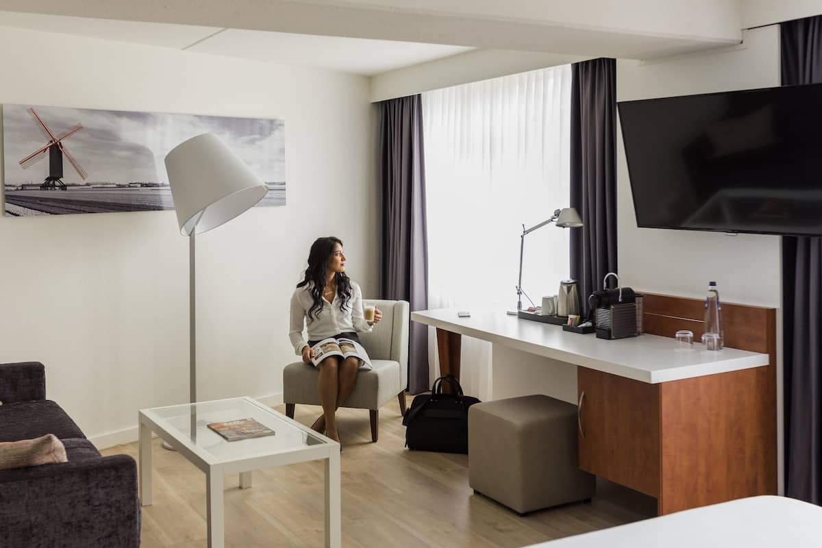 Et moderne hotelværelse med et fladskærms-tv, et skrivebord med lampe, en behagelig lænestol, et sofabord og et maleri af en vindmølle.