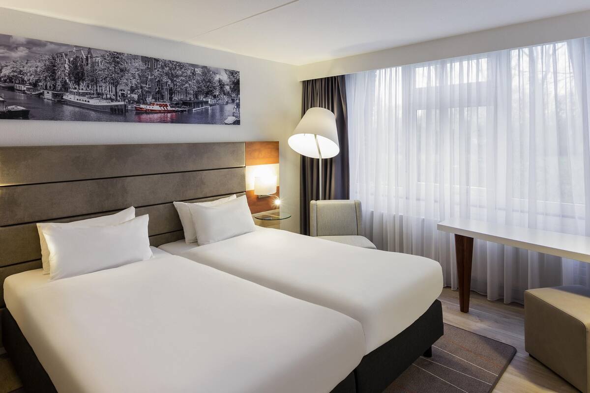 Quarto de hotel com duas camas, uma mesa de cabeceira com abajur, uma mesa e uma janela grande com cortinas leves.