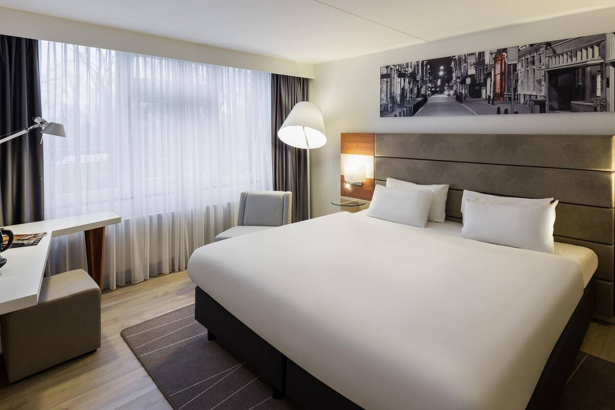 Quarto de hotel com uma cama grande, uma escrivaninha, uma cadeira, um abajur e um quadro na parede.