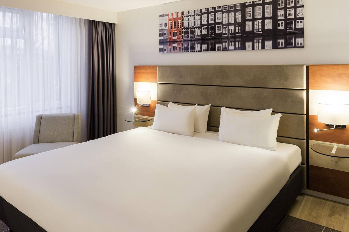 Quarto de hotel com uma cama grande, duas mesas de cabeceira com luminárias, uma cadeira e uma pintura de paisagem urbana na parede.