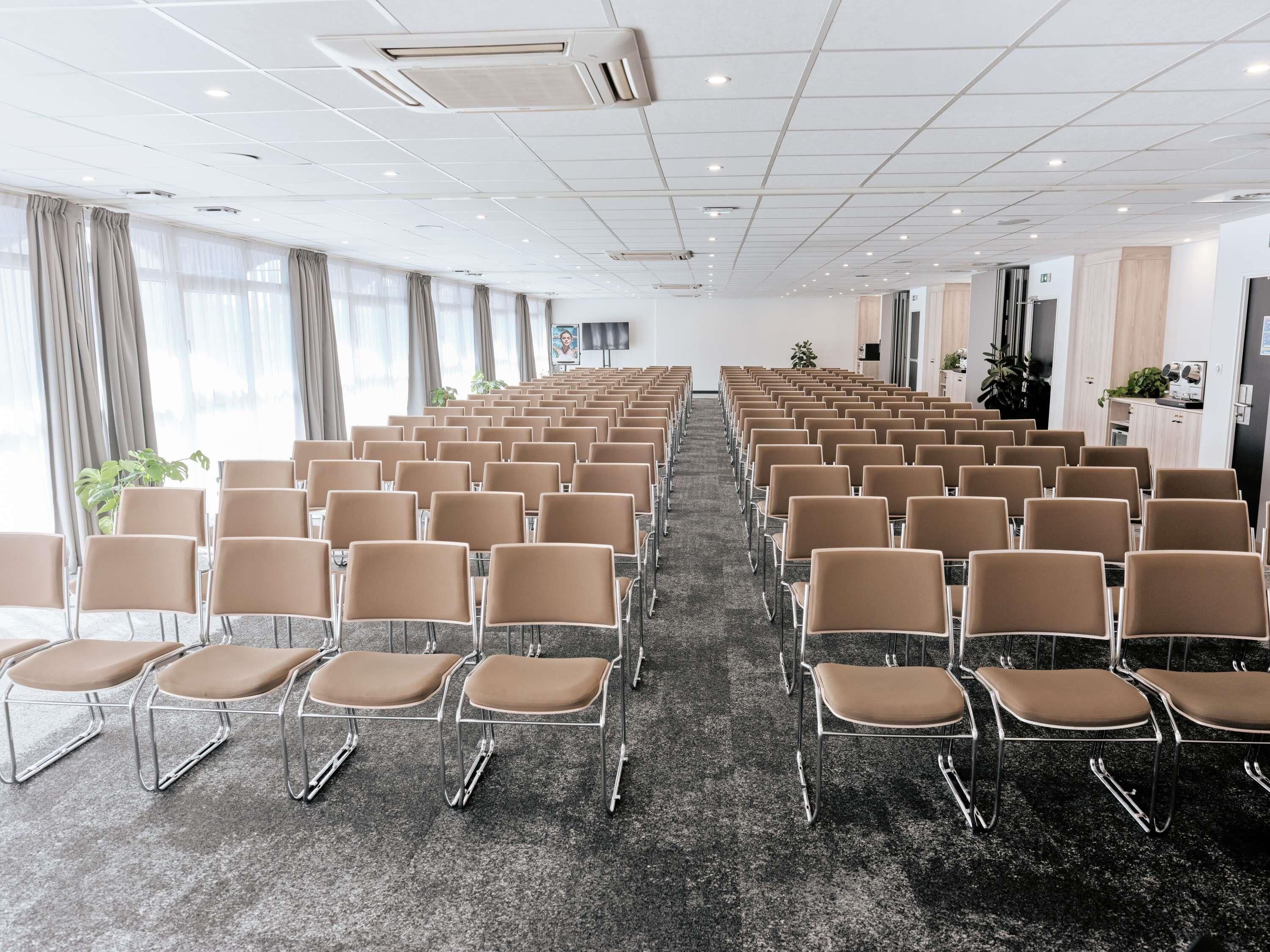 Een vergaderzaal met rijen stoelen, een vloerbedekking en een wit plafond met inbouwverlichting.