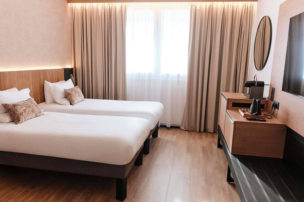 A hotel room with two beds, a wooden dresser, a TV, and a large window with curtains.