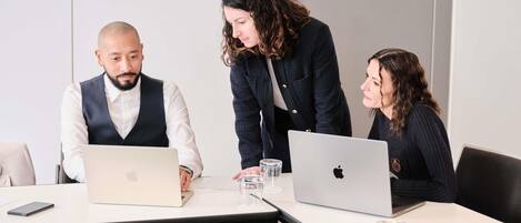 Three people in a meeting room with laptops and glasses of water on the table.