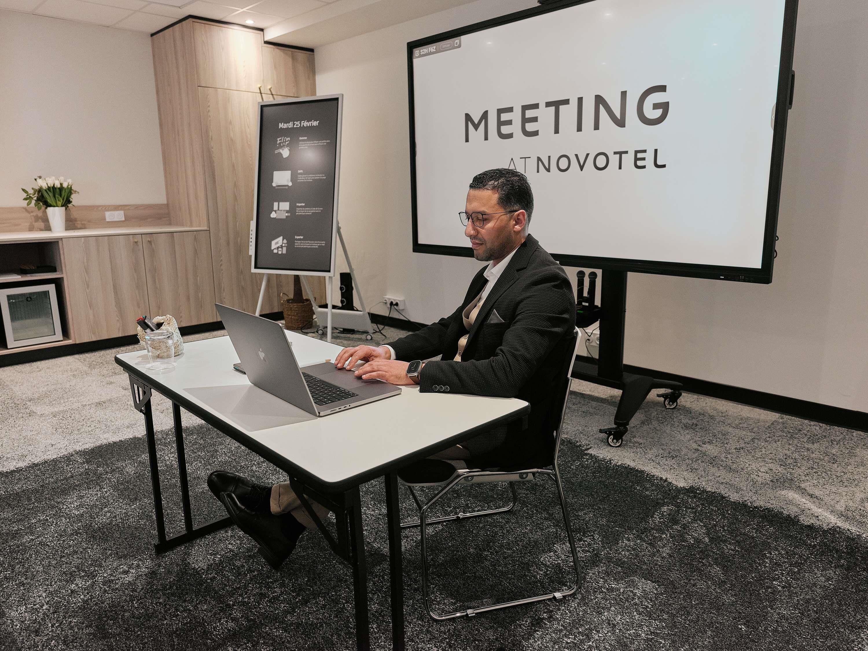 Un home treballant amb un portàtil en una sala de reunions, amb una pantalla de presentació que mostra 'MEETING AT NOVOTEL'.