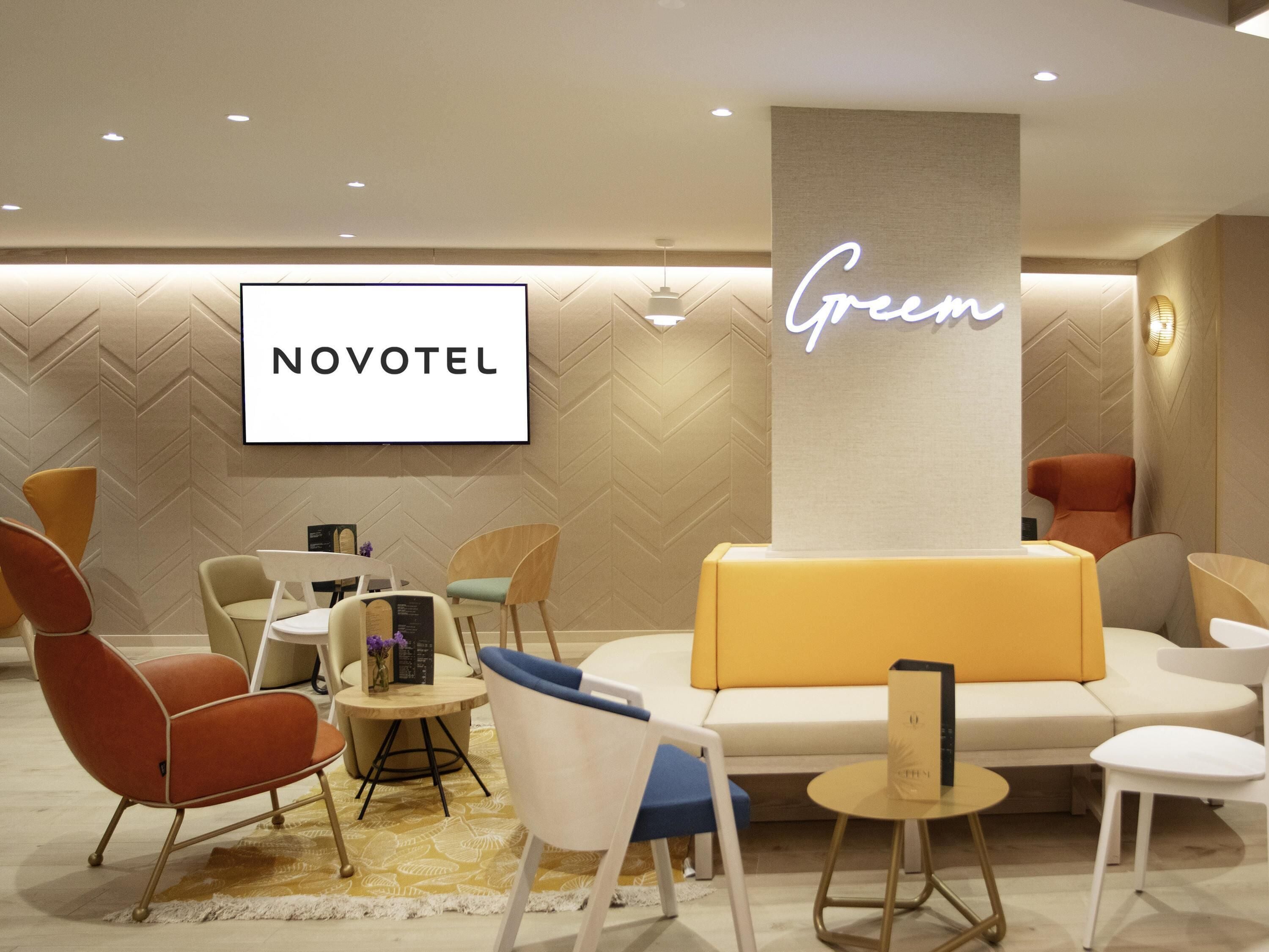 En modern hotellobby med en vägg som har ett fiskbensmönster, en central sittgrupp med en gul soffa och en skylt med texten ”NOVOTEL” ovanför en platt-TV.