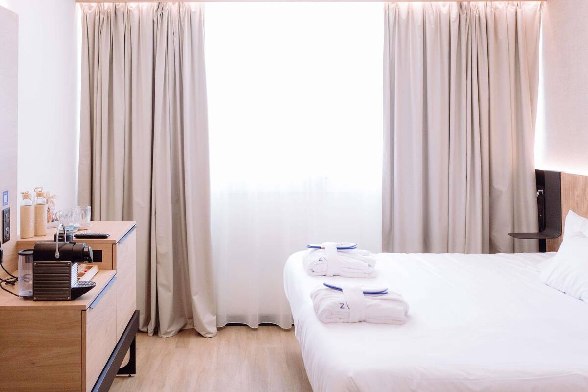 A hotel room with a bed, two towels, a desk with a coffee machine, and a TV.