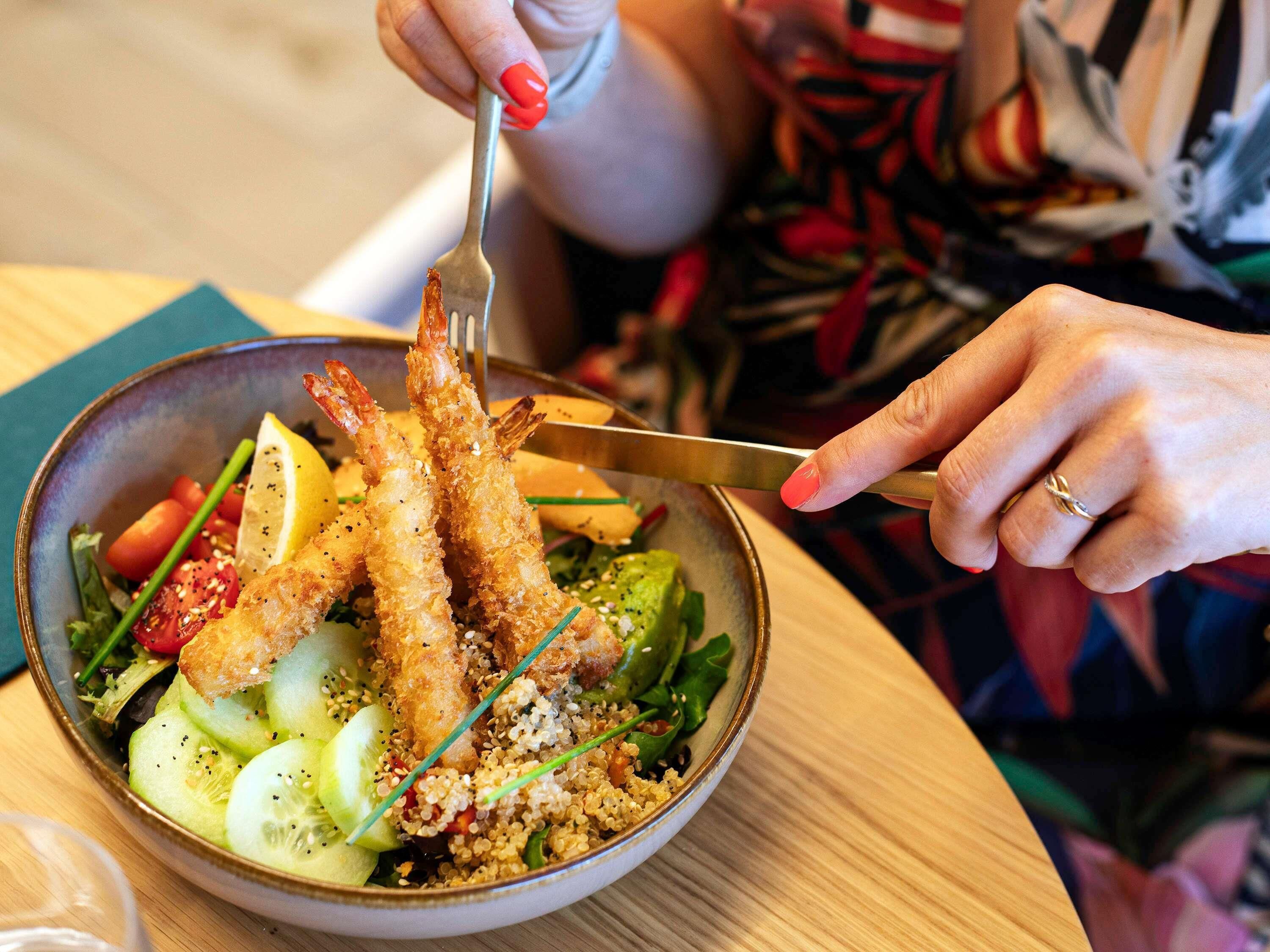 Une personne tenant une fourchette devant un bol contenant des crevettes, une salade et du quinoa.