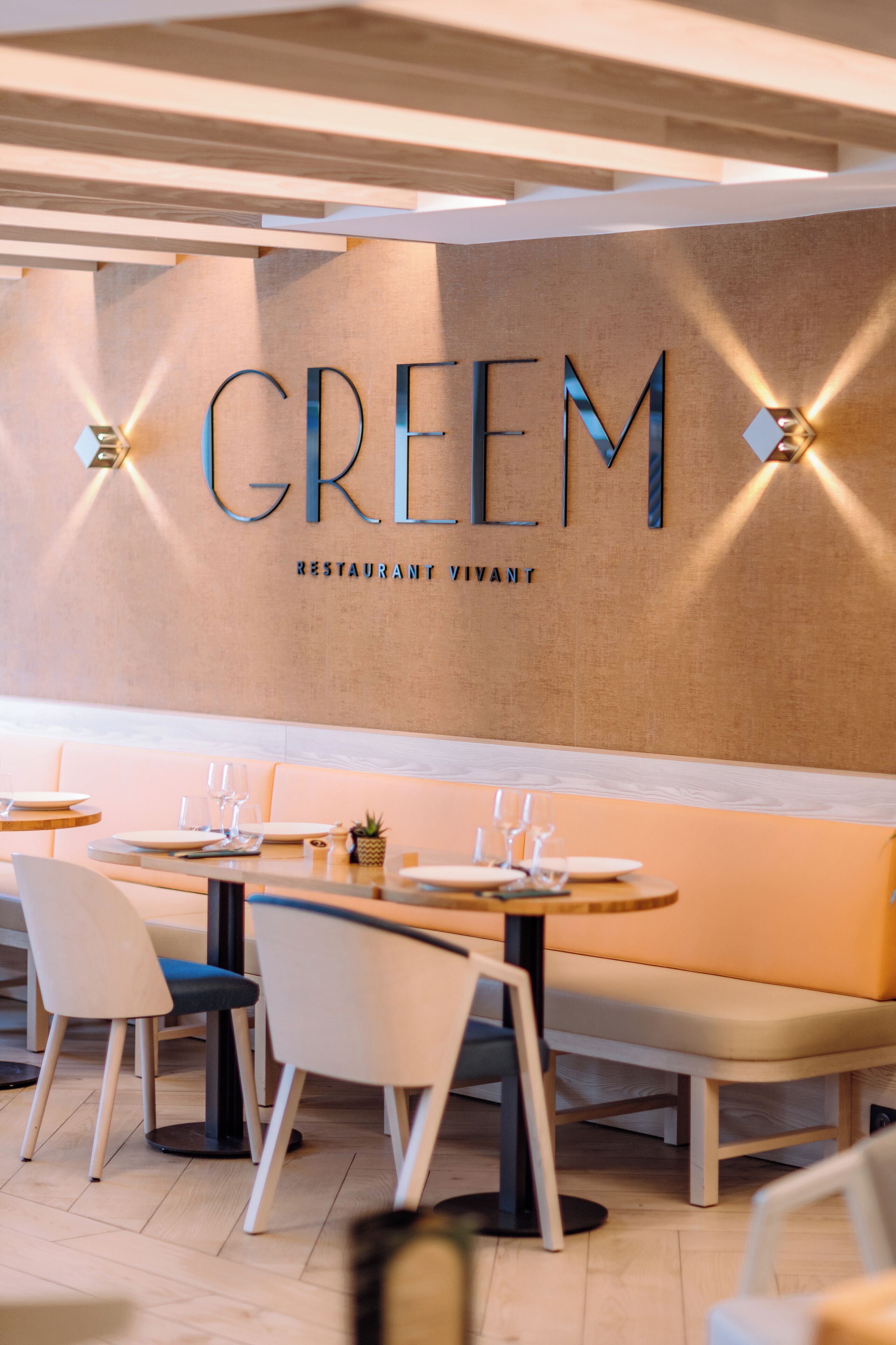 L’intérieur d’un restaurant au design moderne, avec une enseigne fixée au mur sur laquelle on peut lire « GREEM RESTAURANT ». Les tables sont dressées avec des assiettes, des verres et des couverts, et il y a des coins salons avec des sièges rembourrés.