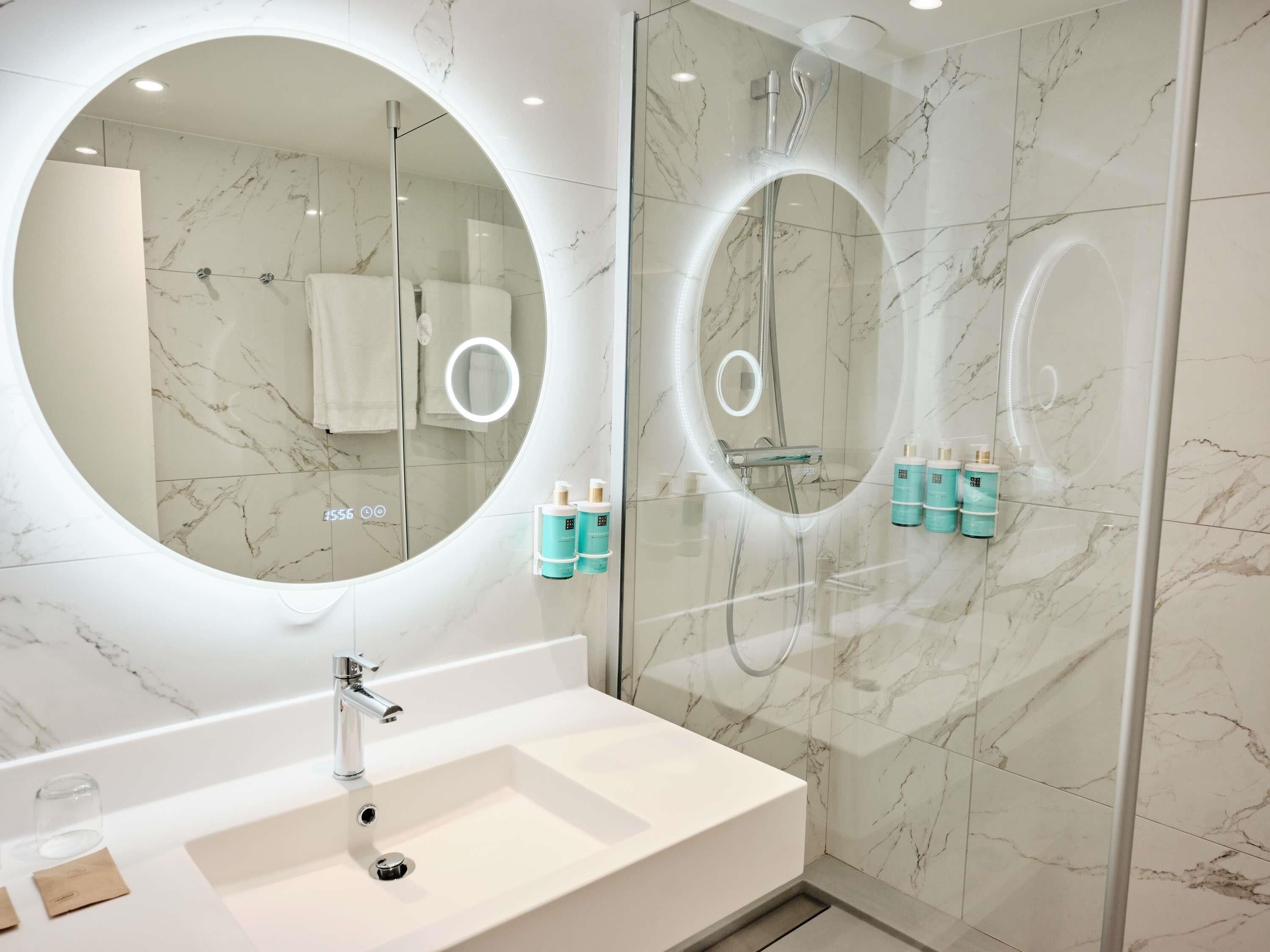 A modern bathroom with a large round mirror, a sink, and a glass-enclosed shower area.