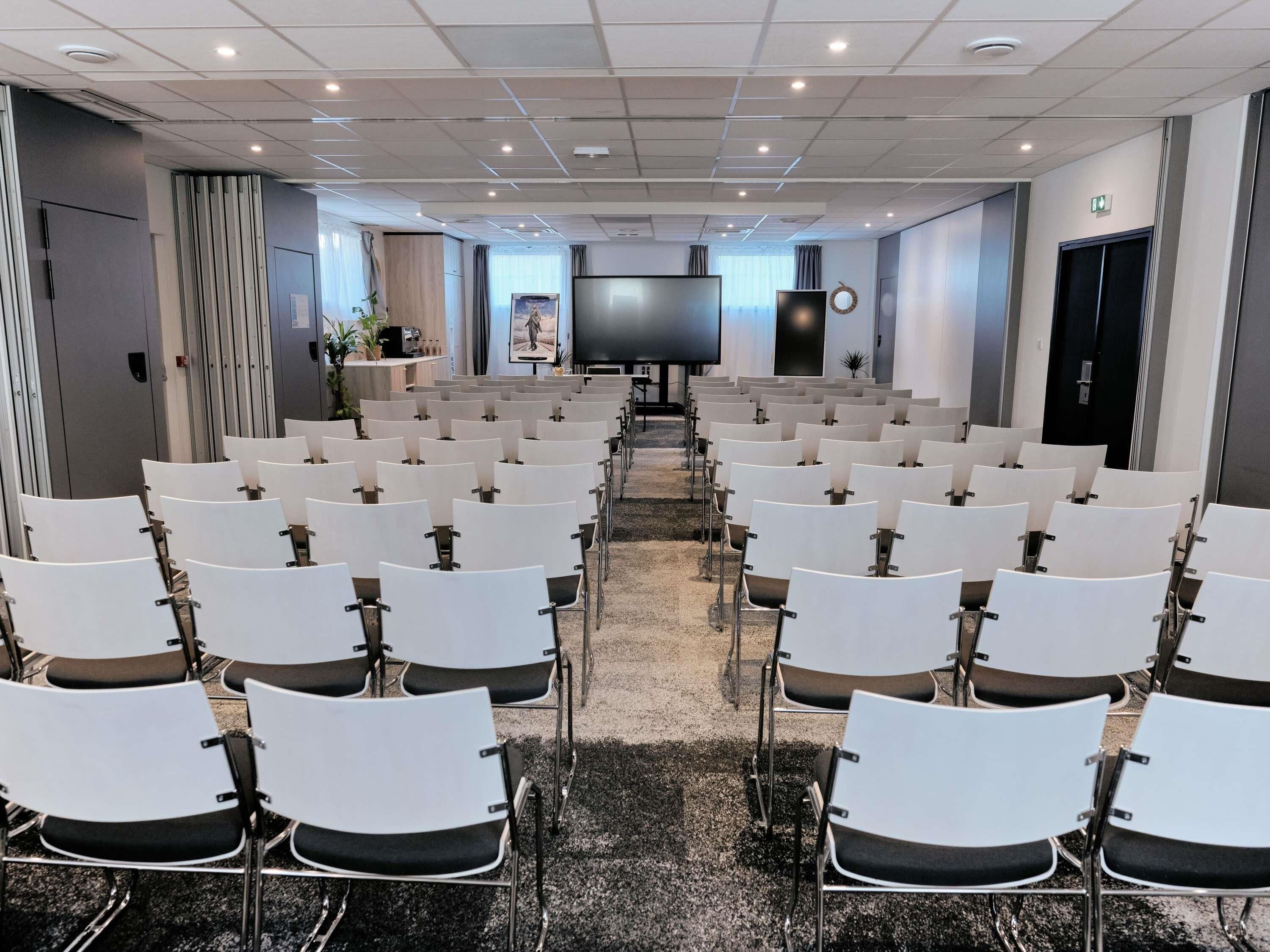 Un salón de conferencias con filas de sillas blancas, una pantalla grande y un proyector.