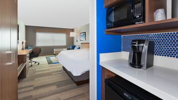 Quarto de hotel com cama, escrivaninha, cadeira, cozinha compacta e janela com persianas.
