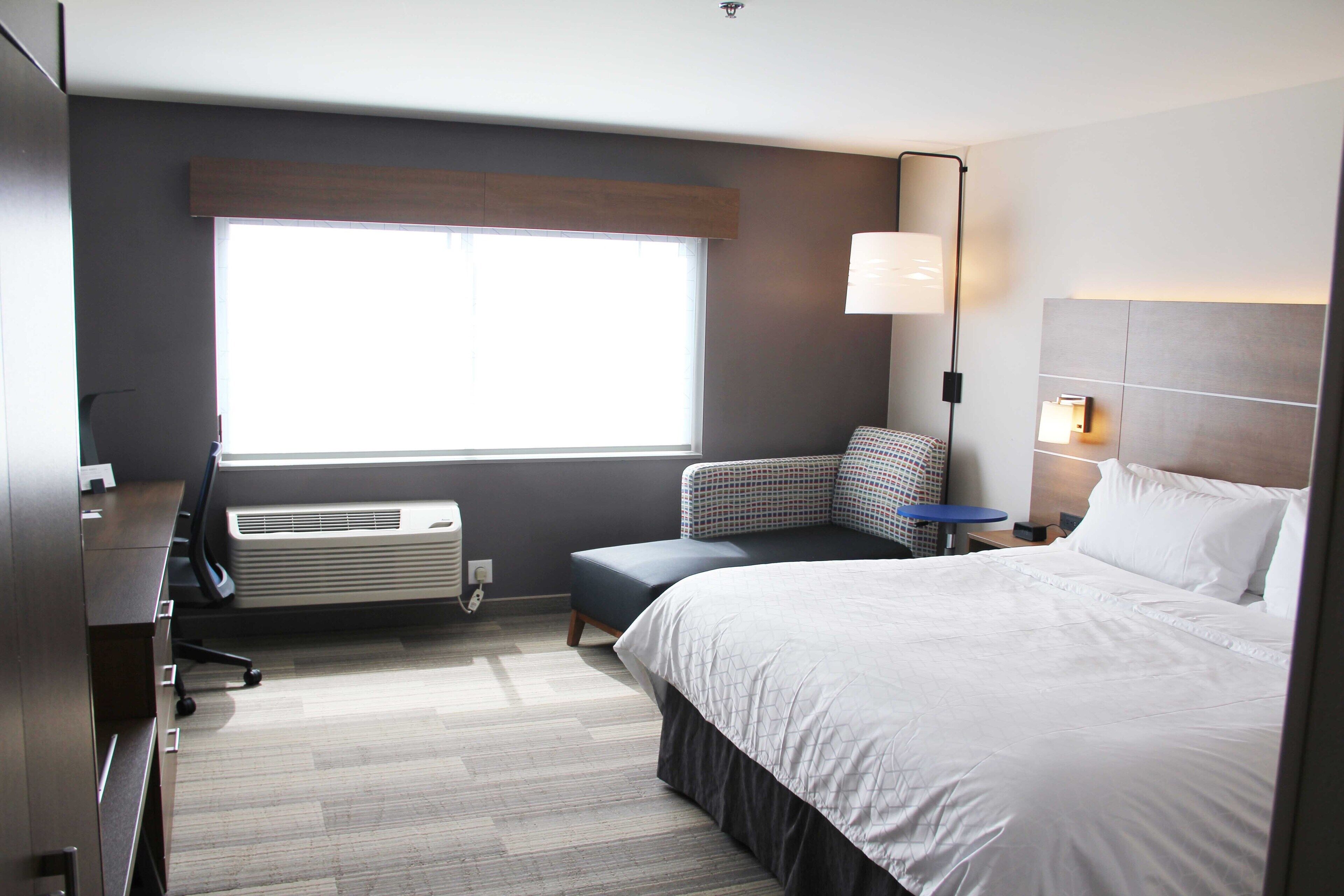 A modern hotel room with a large bed, a desk, a chair, a small table, a lamp, and a window with blinds.