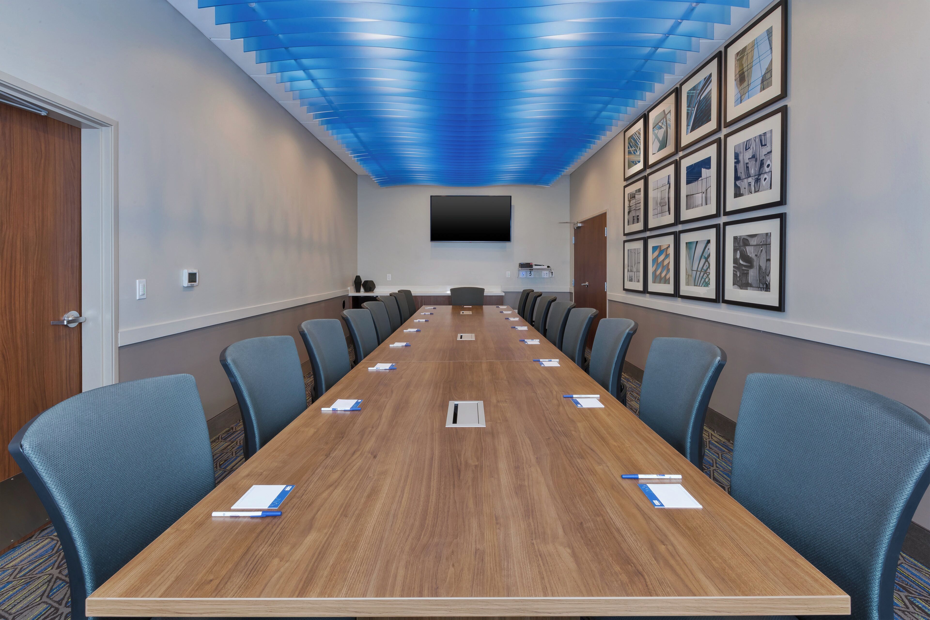 Un salón de conferencias con una mesa larga, sillas azules, un televisor de pantalla plana y obras de arte enmarcadas en la pared.