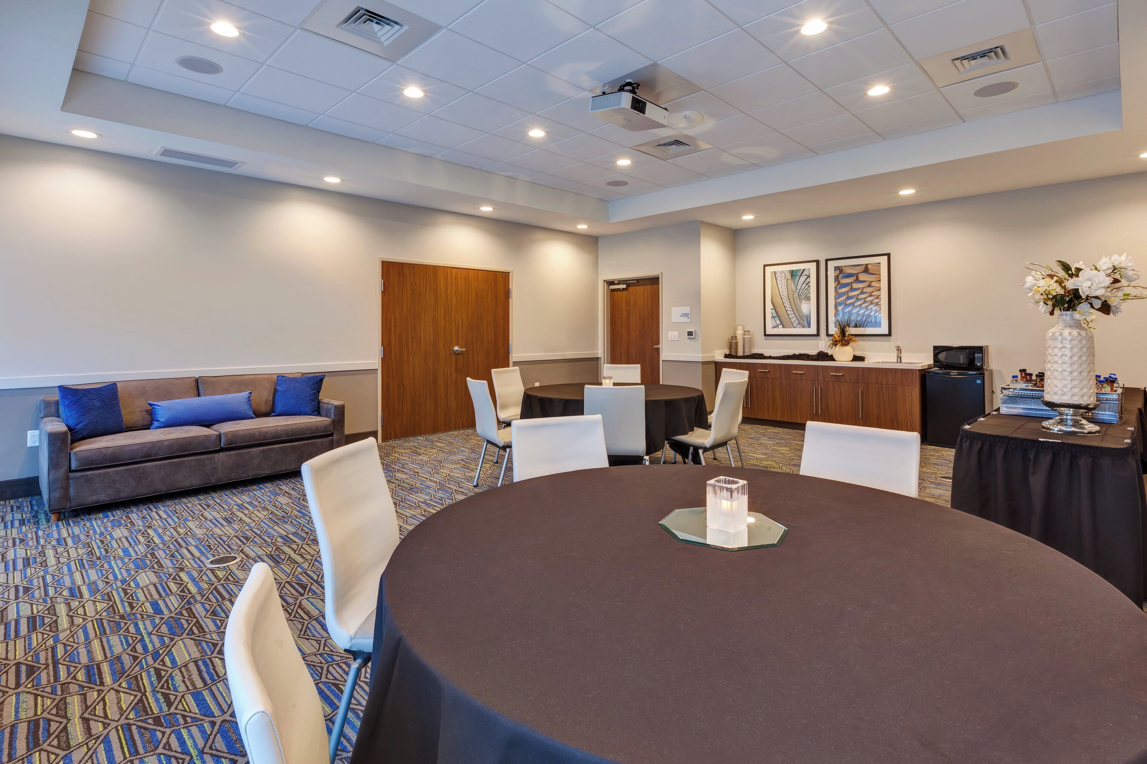 A conference room with a round table, white chairs, a sofa with blue cushions, and a kitchenette area.