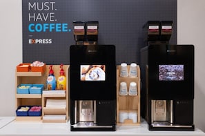 Deux machines à café dotées d’écrans numériques, un distributeur de café et une étagère contenant des dosettes de café et une cafetière.